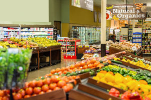 Colorful fresh fruits and vegetables are displayed in produce section of local grocery store or supermarket. Rows of tomatoes, peppers, potatoes, bananas, apples, and other natural organic produce is arranged on shelves and tables.