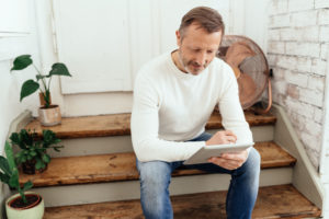 Athletic casual middle-aged man sitting on wooden interior steps in a loft with potted plants reading or watching media on a tablet-pc with copyspace